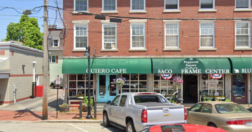 Exterior of Quiero Cafe with green awning, surrounded by parked cars and nearby shops in a quaint town square.