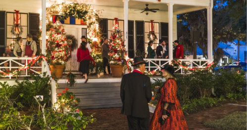 A festive scene with people in holiday attire at a decorated porch, adorned with lights and greenery.