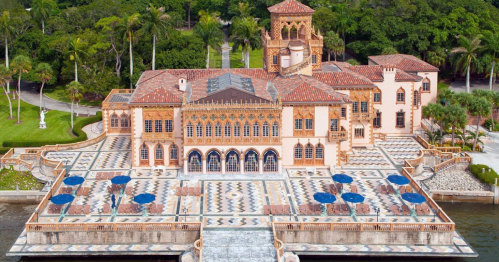 Aerial view of a grand, ornate mansion with a tiled terrace, surrounded by lush greenery and palm trees.