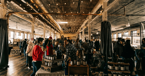 A bustling indoor market with people browsing stalls, string lights overhead, and a warm, inviting atmosphere.