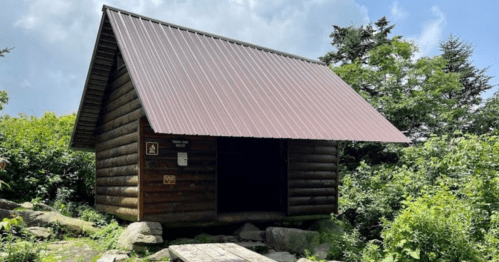 A small wooden cabin with a metal roof, surrounded by greenery and under a partly cloudy sky.