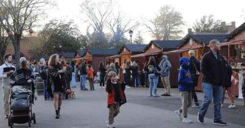 A bustling outdoor market scene with people walking, shopping, and socializing near festive stalls.