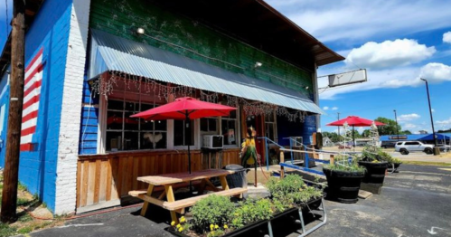 Colorful building with a blue and green exterior, red umbrellas on picnic tables, and a small American flag painted on the wall.