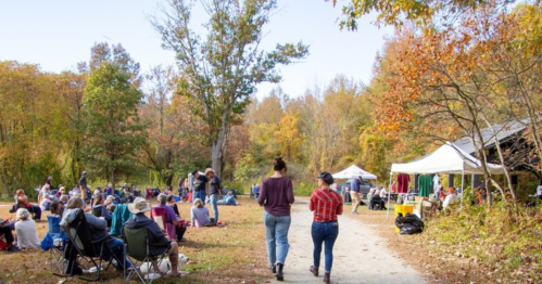 Two people walk along a path in a vibrant autumn park filled with people enjoying a gathering under colorful trees.