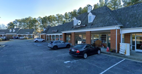 A row of shops with a parking lot, featuring brick buildings and a tree-lined background.