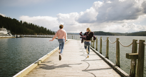 Two people run joyfully along a wooden pier by a lake, surrounded by trees and a cloudy sky.