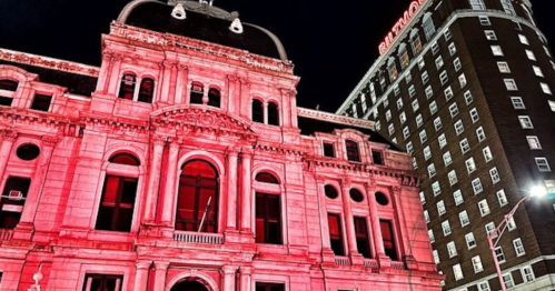 Historic building illuminated in red at night, with a tall hotel in the background.