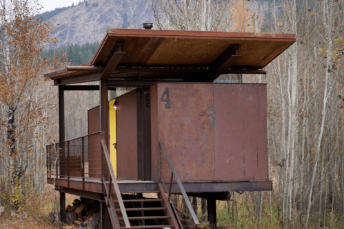 A modern cabin with a rust-colored exterior, elevated on stilts, surrounded by trees and mountains in the background.