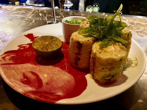 Grilled corn on the cob with green onions, served with a lime wedge on a decorative plate.