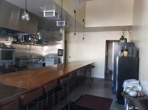 A modern kitchen with stainless steel appliances, a long wooden counter, and bar stools, featuring pendant lighting.