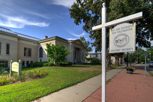 Sign marking the entrance to the Colchester Historic District, with a historic building and greenery in the background.