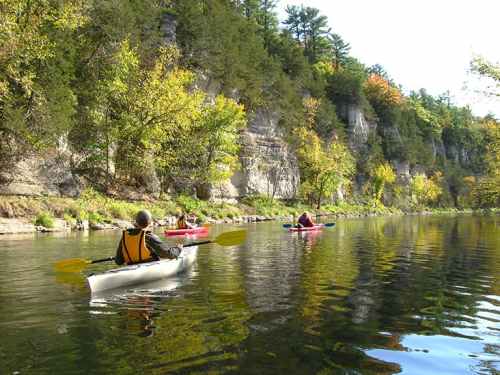 Two kayakers paddle on a calm river surrounded by lush green trees and rocky cliffs under a clear blue sky.