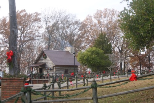 A rustic cabin decorated with red bows, surrounded by autumn trees and a wooden fence. Smoke rises from the chimney.