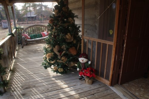 A cozy porch with a decorated Christmas tree and a snowman figure beside a potted plant, surrounded by wooden elements.