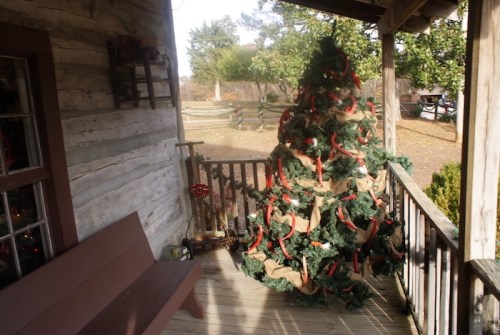 A rustic porch with a decorated Christmas tree and a wooden bench, surrounded by greenery and a wooden fence.
