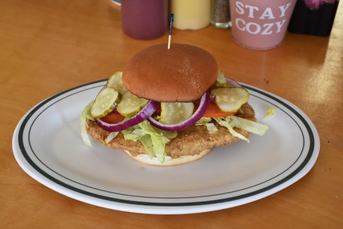 A crispy chicken sandwich topped with pickles, lettuce, tomatoes, and onions on a bun, served on a white plate.