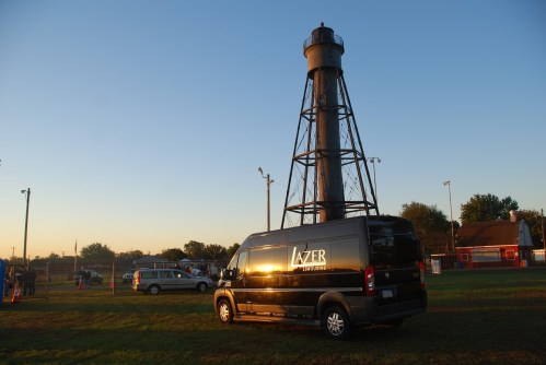 A black van parked near a tall, vintage water tower at sunset, with a clear sky and grassy field in the background.