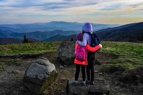 Two girls embrace while overlooking a scenic mountain view, with rolling hills and a colorful sky in the background.