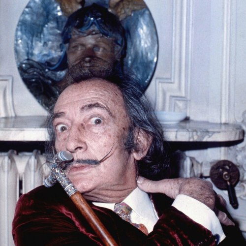 A man with a prominent mustache and cane poses dramatically, with a decorative wall piece in the background.