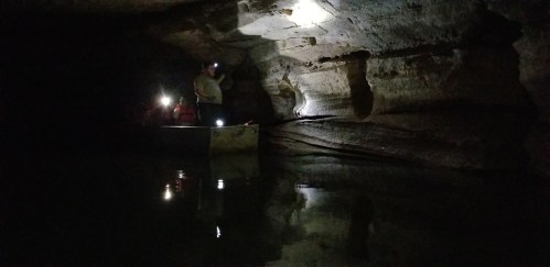 A person stands on a small boat in a dark cave, illuminated by headlamps, with water reflecting the rocky walls.