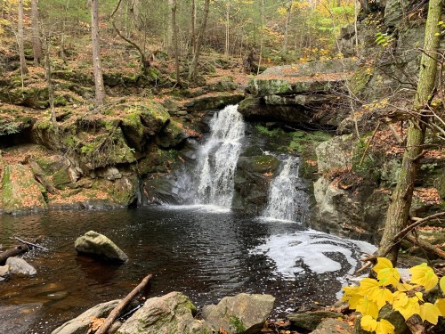 A serene waterfall cascades into a dark pool, surrounded by autumn foliage and rocky terrain in a forested area.