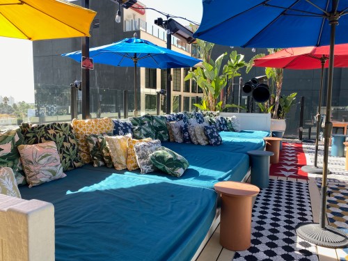 A vibrant rooftop lounge with colorful umbrellas, plush seating, and decorative pillows surrounded by greenery.
