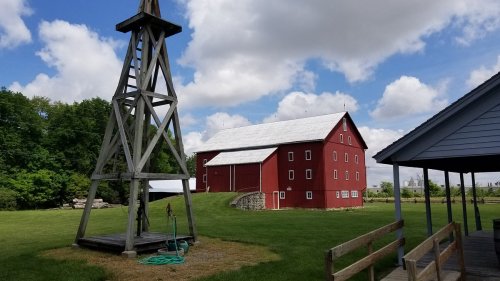 A red barn stands beside a wooden windmill under a blue sky with fluffy clouds, surrounded by green grass.