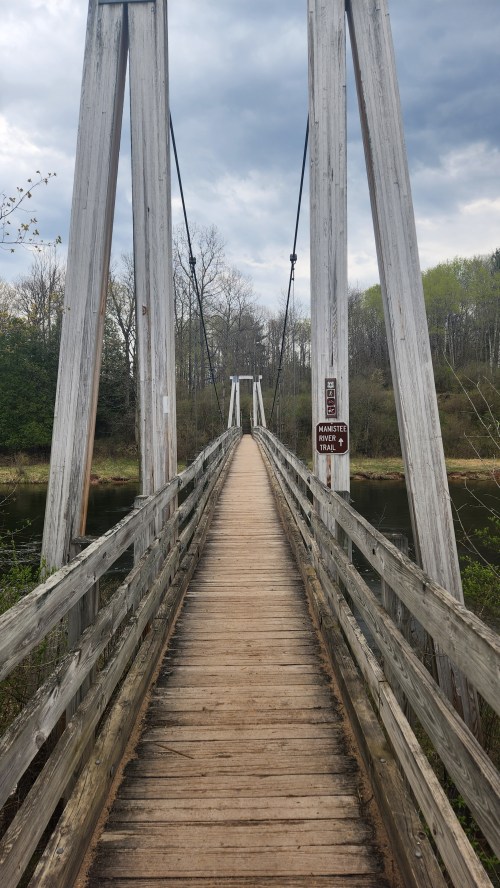 A wooden suspension bridge stretches over a calm body of water, surrounded by trees and a cloudy sky.