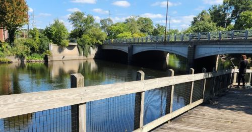 A serene river scene with a bridge, lush greenery, and a wooden walkway along the water's edge under a clear blue sky.