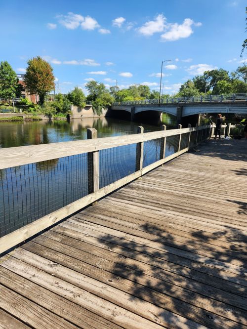 A wooden walkway by a calm river, with a bridge in the background and trees under a blue sky.