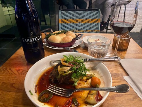 A plate of beef with vegetables, a bottle of Saratoga sparkling water, a glass of red wine, and bread rolls on a wooden table.