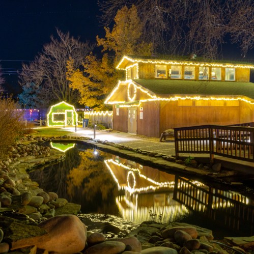 A cozy building adorned with festive lights reflects in a calm waterway at night, surrounded by autumn trees.