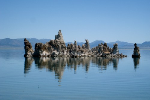 A rocky formation rises from calm water, reflecting in the serene lake with mountains in the background under a clear blue sky.