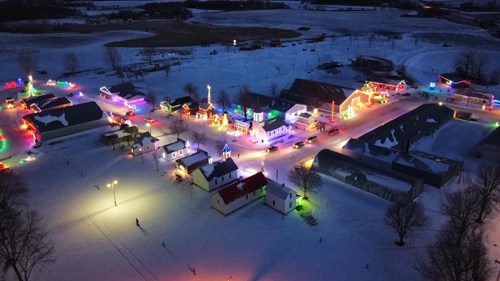 Aerial view of a snowy village illuminated with colorful holiday lights at night.