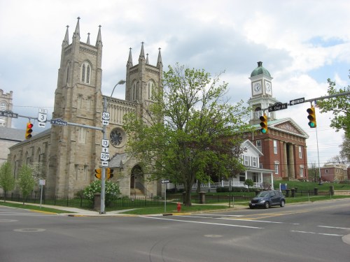 Historic church with twin spires and a clock tower, set at a street intersection with traffic lights and greenery.