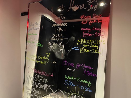 A mirror covered in colorful writing, displaying restaurant hours and events for "Mama's Kitchen."