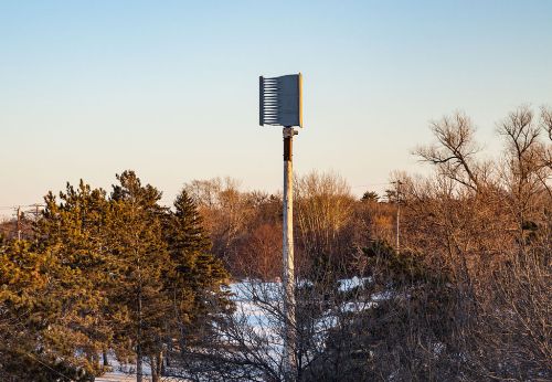 A tall outdoor warning siren on a pole, surrounded by trees and a clear sky during sunset.