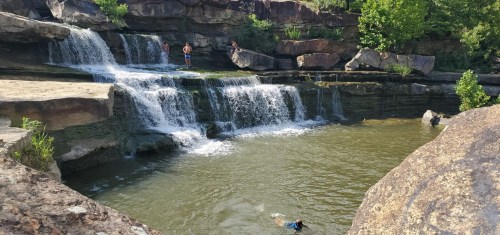 A serene waterfall cascades into a calm pool, with people enjoying the natural setting and swimming nearby.