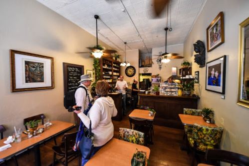 A cozy café interior with wooden tables, floral chairs, and patrons chatting near the counter. Art adorns the walls.