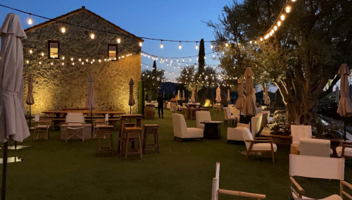 A cozy outdoor restaurant at dusk, with string lights illuminating tables and chairs surrounded by greenery.