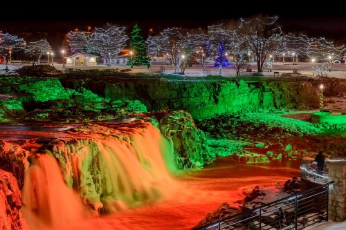 A waterfall illuminated in red and green lights, surrounded by snowy trees and festive decorations at night.