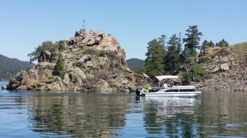 A boat approaches a rocky island with trees and an American flag on a clear, sunny day. Reflections shimmer on the water.
