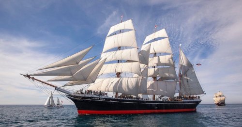 A majestic tall ship with white sails gliding through calm waters under a blue sky, with another ship in the background.