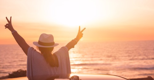 A person in a hat raises their arms in peace signs, enjoying a sunset over the ocean from a car rooftop.