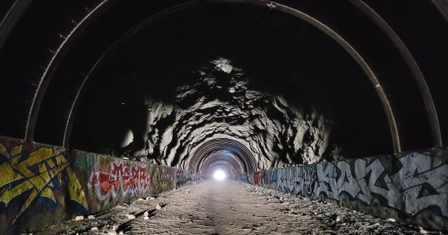 A dark tunnel with colorful graffiti on the walls, leading to a bright light at the far end.