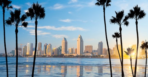 City skyline at sunset with palm trees in the foreground and reflections on the water.