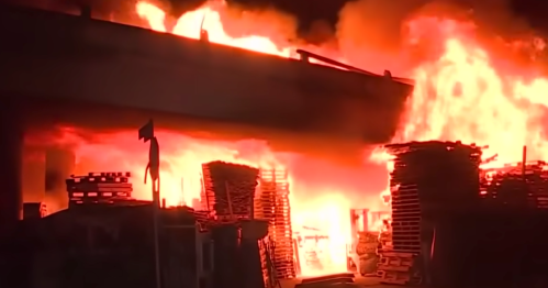 A large fire engulfing a structure with wooden pallets, creating intense flames and smoke at night.