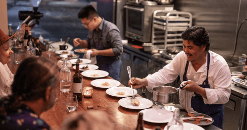 A chef serves dishes at a bar while a second cook prepares food in a bustling kitchen setting. Guests watch eagerly.
