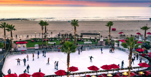 A beachfront ice skating rink at sunset, surrounded by palm trees and festive red umbrellas, with people skating and enjoying the view.