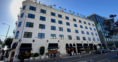 A white, multi-story building with large windows and a rooftop garden, located on a city street.